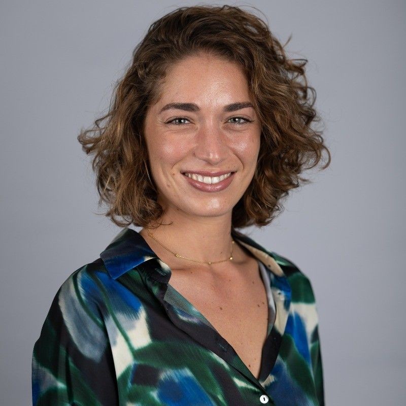 Smiling person with curly hair wearing a colorful patterned shirt against a plain background.