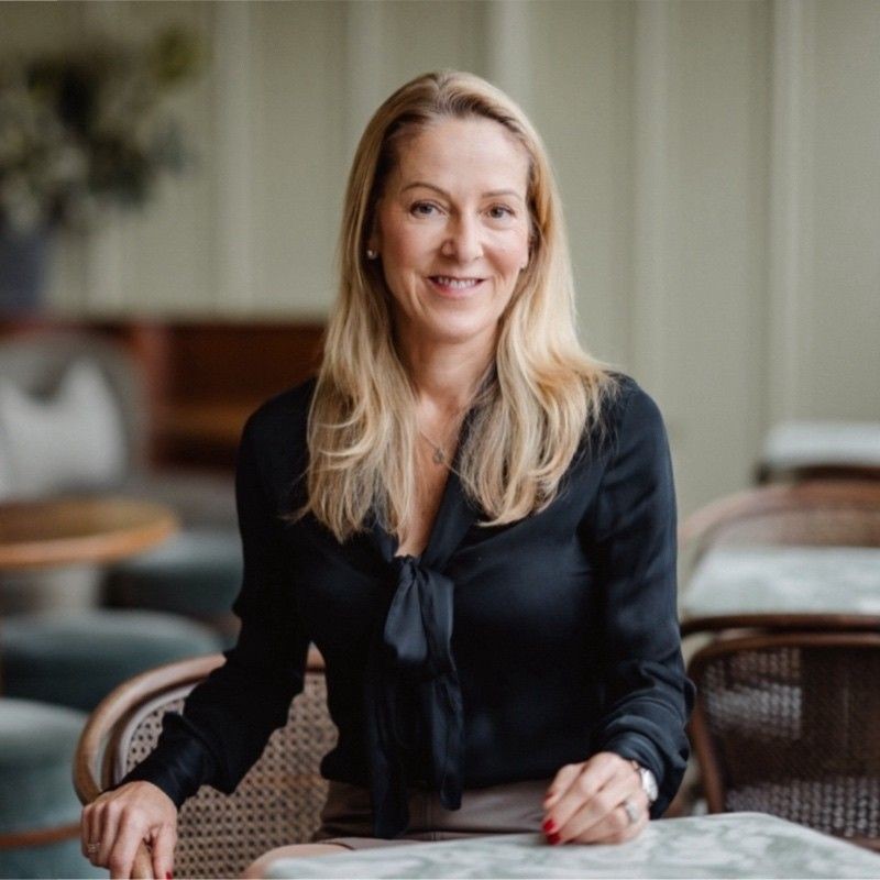 Smiling woman with long blonde hair in a black blouse sitting in a light-colored cafe setting.